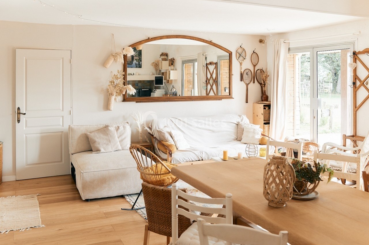 Salon lumineux avec canapé beige, table en bois, chaises blanches, miroir mural et grande baie vitrée donnant sur un jardin.