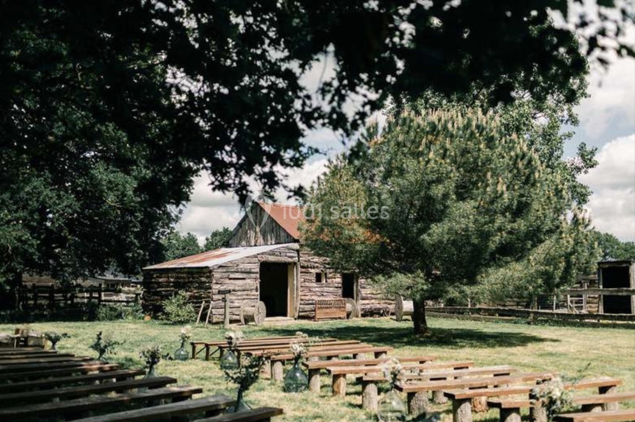 Vieille grange en bois entourée d'arbres, avec des bancs disposés en extérieur sur une pelouse.
