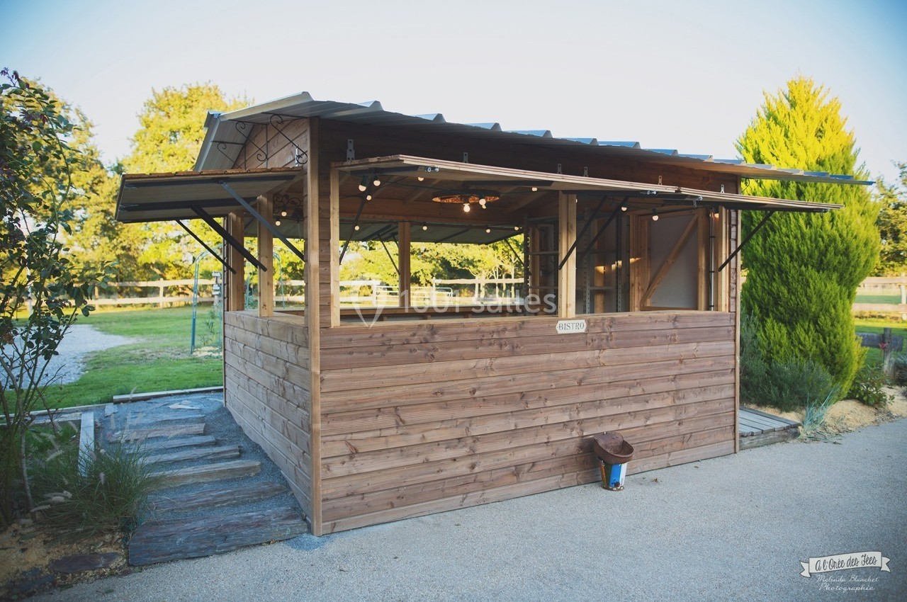Kiosque en bois avec toit en tôle, éclairage suspendu, situé dans un espace extérieur verdoyant.