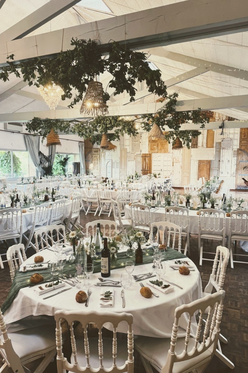 Salle de réception décorée avec des tables rondes dressées, guirlandes végétales suspendues et lumière naturelle.