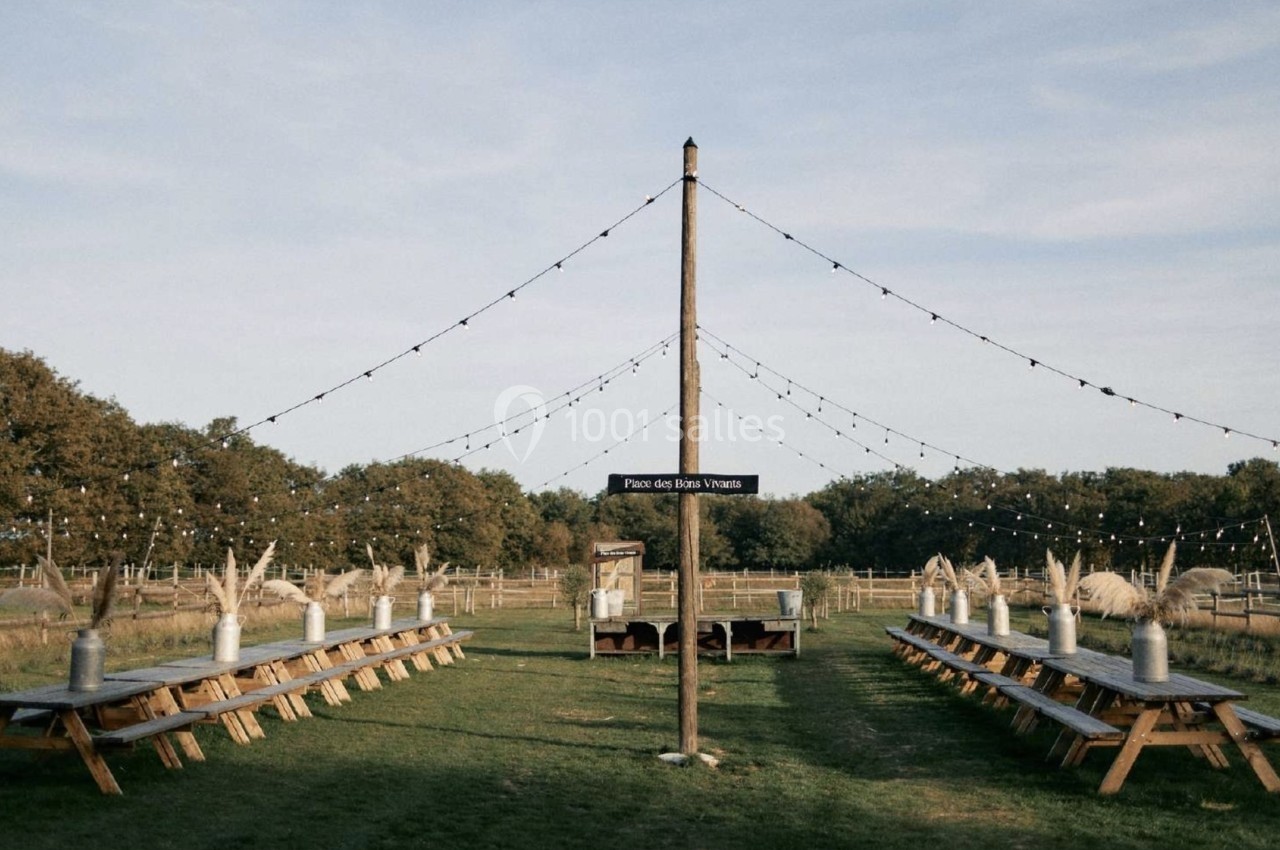 Deux longues tables en bois disposées en extérieur sous des guirlandes lumineuses, entourées de champs et d'arbres.