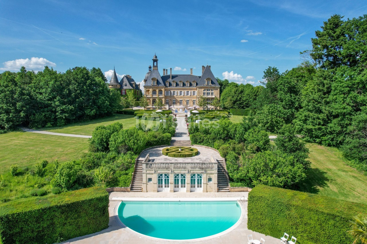 Vue aérienne d'un château entouré de jardins paysagers, avec une piscine au premier plan et des arbres en arrière-plan.