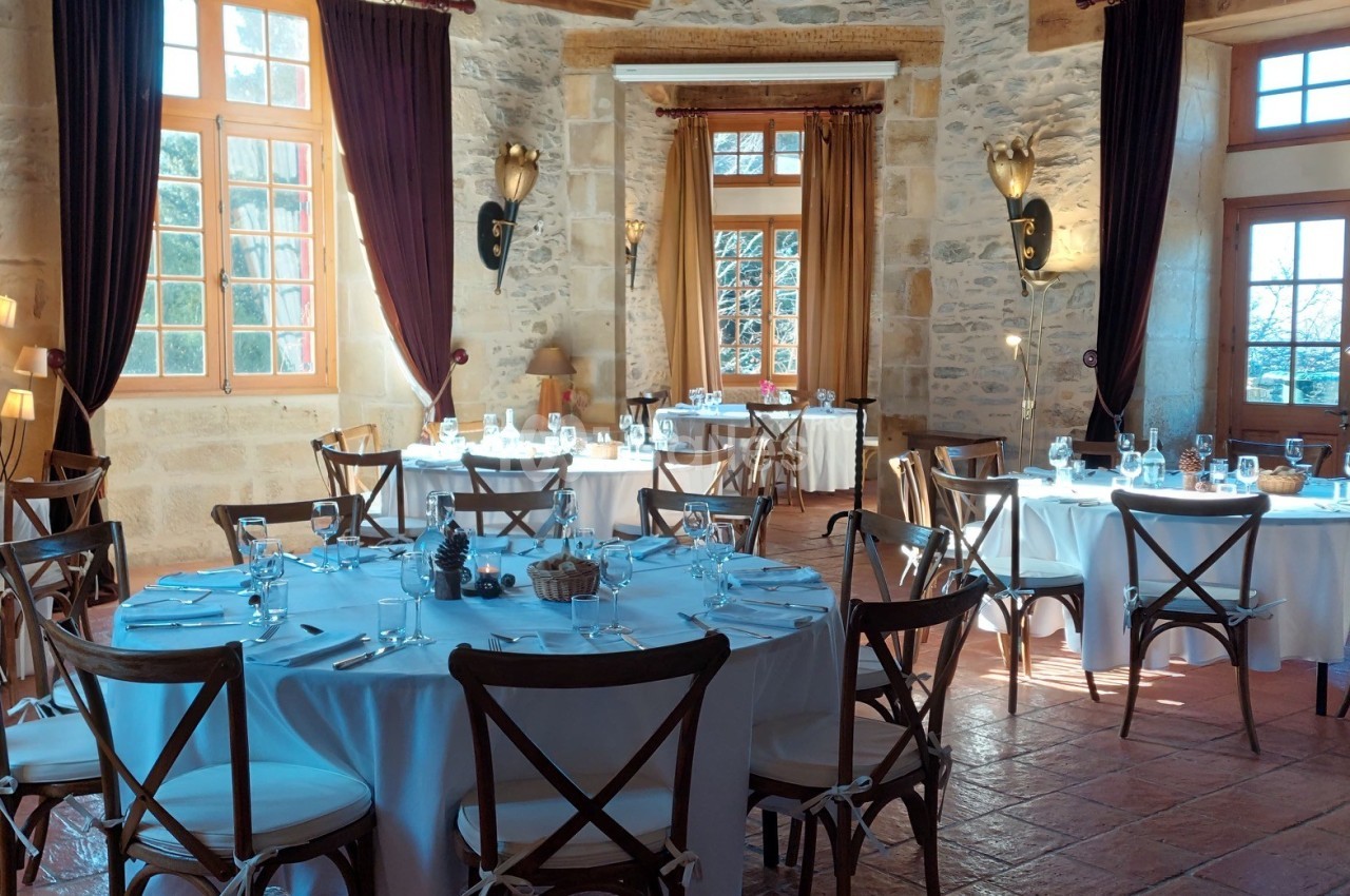 Salle de réception lumineuse avec tables rondes dressées, chaises en bois et murs en pierre apparente.
