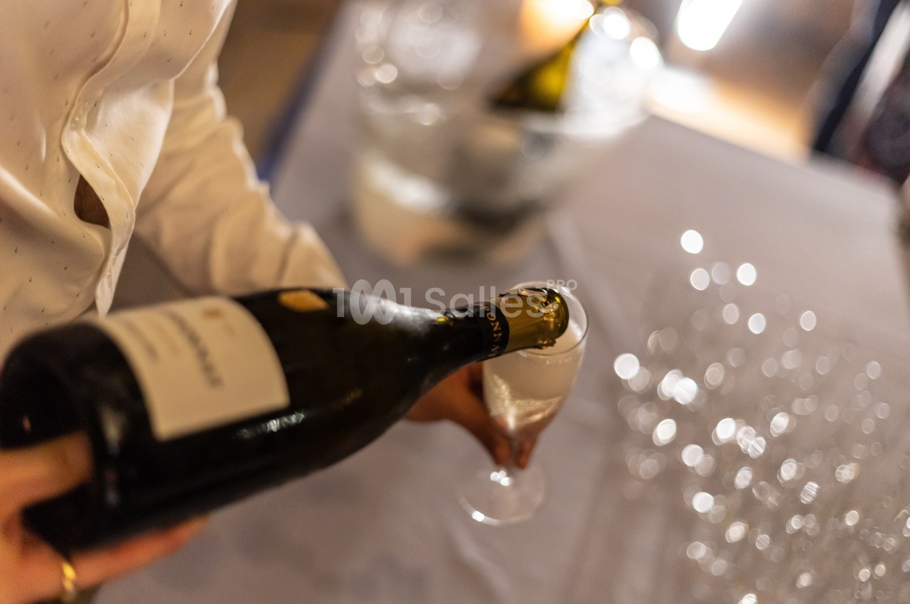 Une personne verse du vin mousseux dans une flûte sur une table avec des verres et un seau à glace en arrière-plan.