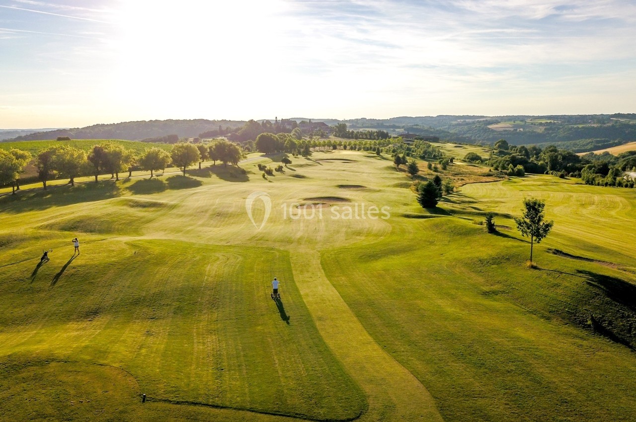 Vue aérienne d'un terrain de golf verdoyant avec des arbres alignés et un paysage vallonné sous un ciel ensoleillé.