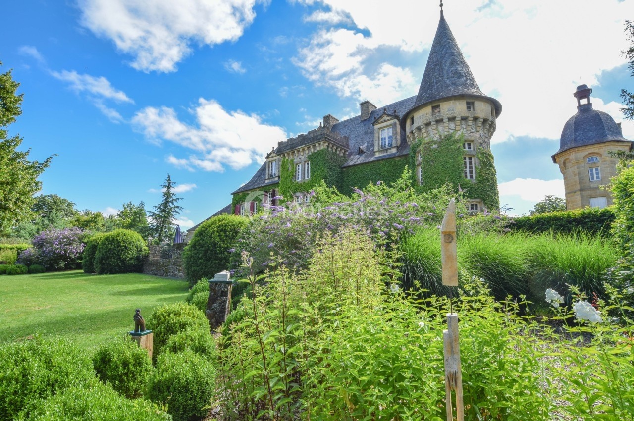 Château ancien entouré de jardins verdoyants, avec une tour en pierre et un ciel partiellement nuageux.