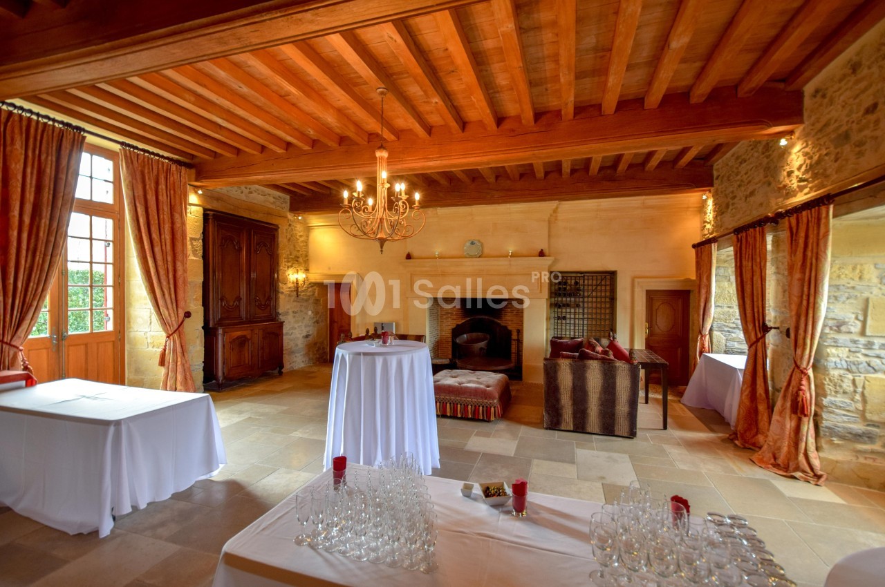 Salle de réception avec poutres apparentes, cheminée en pierre, tables dressées et grandes fenêtres avec rideaux.