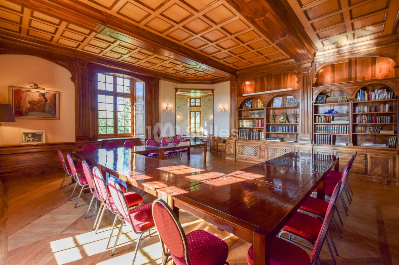 Salle de réunion lumineuse avec une grande table en bois, des chaises rouges et des étagères remplies de livres.