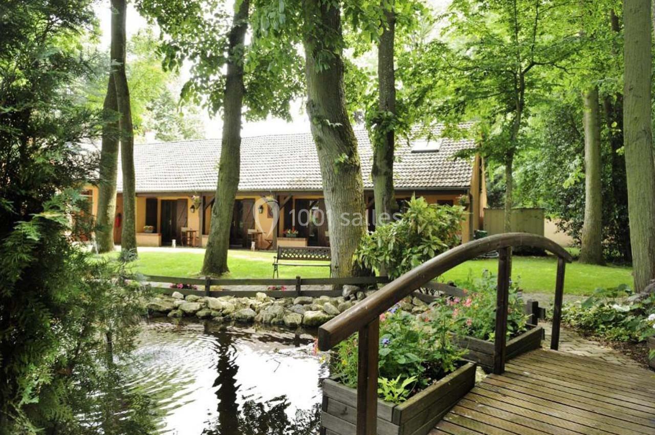 Location salle Les Villages Vovéens (Eure-et-Loir) - Hôtel Restaurant le Quai Fleuri #4 Maison entourée d'arbres avec un jardin, un étang et un petit pont en bois au premier plan.