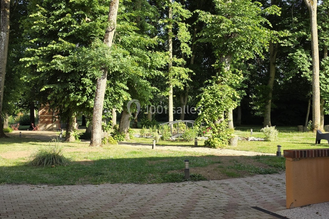 Location salle Les Villages Vovéens (Eure-et-Loir) - Hôtel Restaurant le Quai Fleuri #21 Vue d'un jardin arboré avec des arbres, des plantes et des chemins pavés sous une lumière ensoleillée.