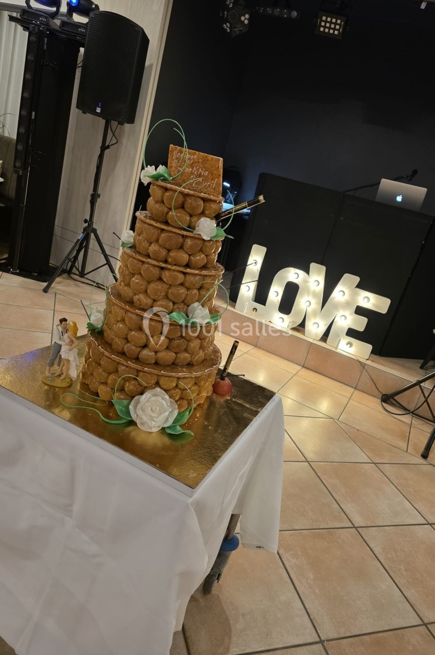 Pièce montée en choux décorée de fleurs et d'une figurine de mariage, posée sur une table devant une décoration lumineuse…