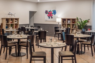 Salle de restaurant lumineuse avec tables dressées, chaises en bois sombre, étagères décoratives et plantes vertes.