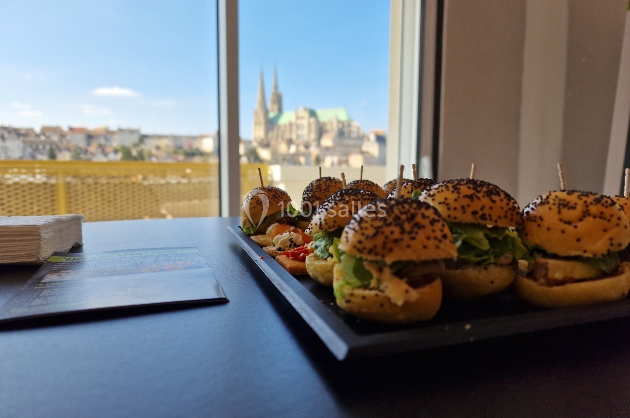 Plateau de mini-burgers garnis, posé sur une table près d'une fenêtre avec vue sur une ville et une cathédrale.