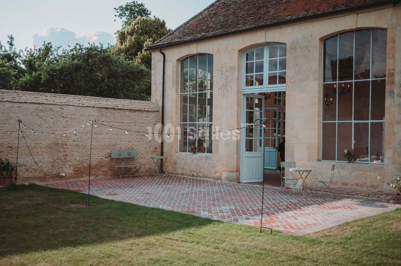 Terrasse pavée devant une bâtisse en pierre avec grandes fenêtres, entourée de verdure et décorée de guirlandes lumineuses.