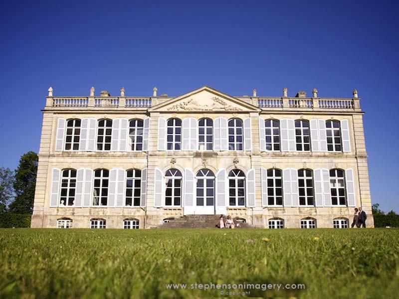 Façade d'un grand bâtiment historique avec de nombreuses fenêtres, entouré d'une pelouse sous un ciel bleu.