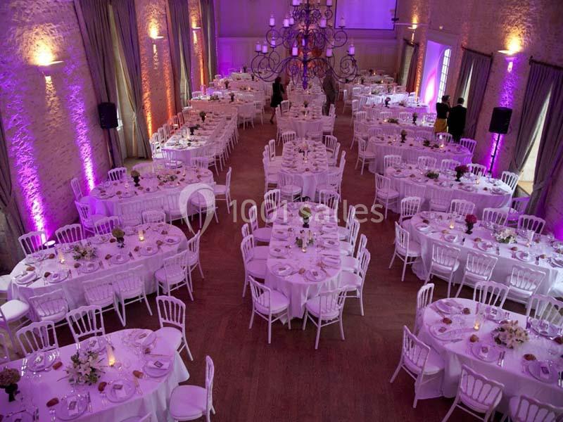 Salle de réception décorée avec des tables rondes, nappes blanches, bouquets de fleurs et éclairage violet.
