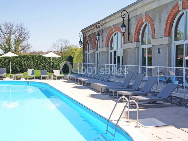 Piscine extérieure bordée de chaises longues grises, parasols blancs et bâtiment avec grandes baies vitrées.
