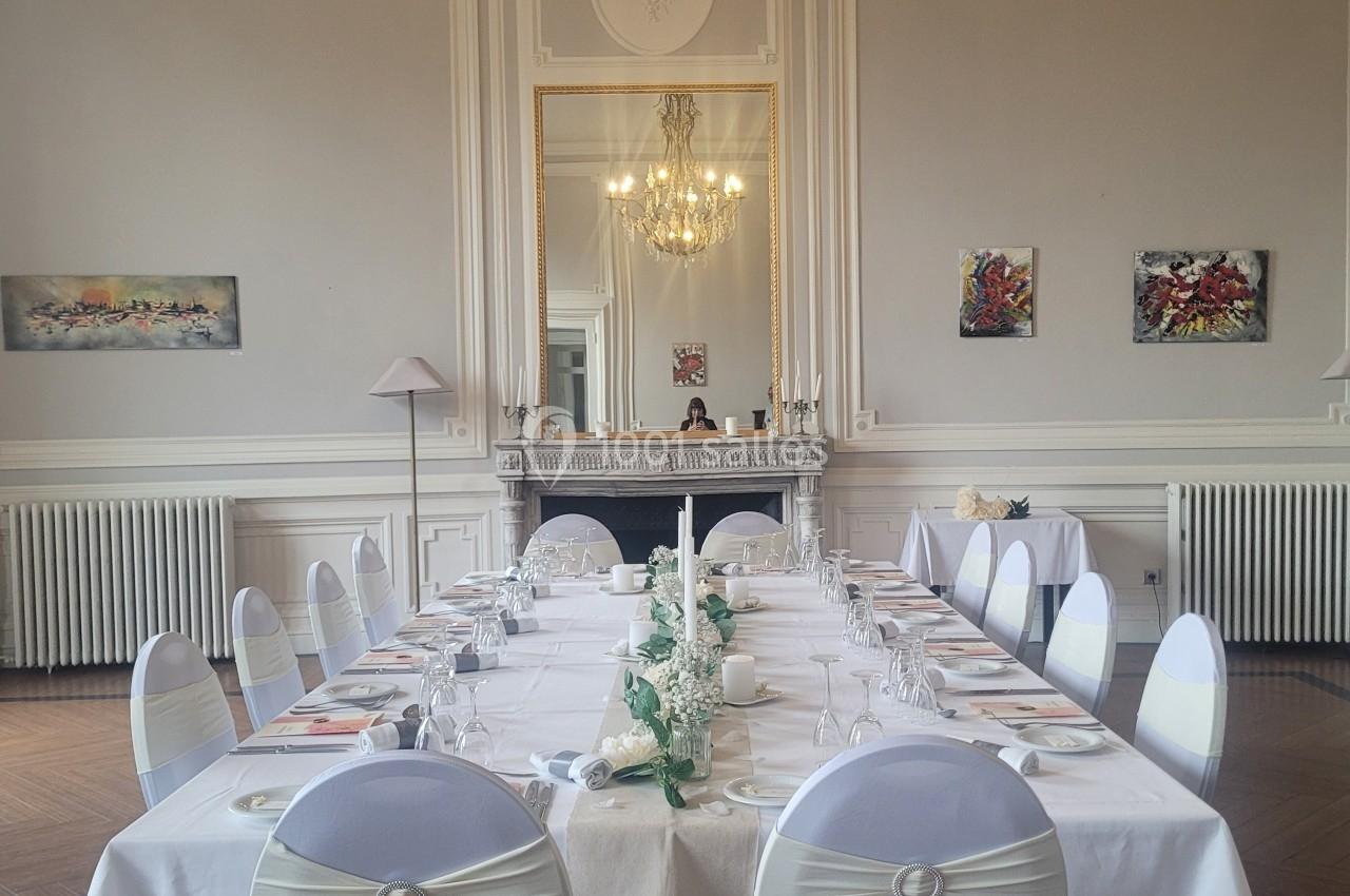 Salle élégante avec grande table dressée, chaises blanches décorées, lustre imposant et miroir encadré sur un mur clair.