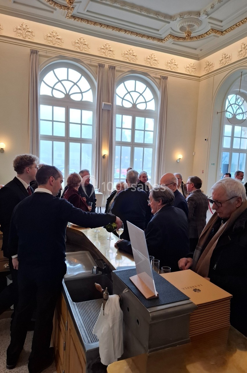 Location salle Contrexéville (Vosges) - Casino Partouche de Contrexéville #21 Un groupe de personnes discute près d'un comptoir dans une salle lumineuse avec de grandes fenêtres et un plafond orné.