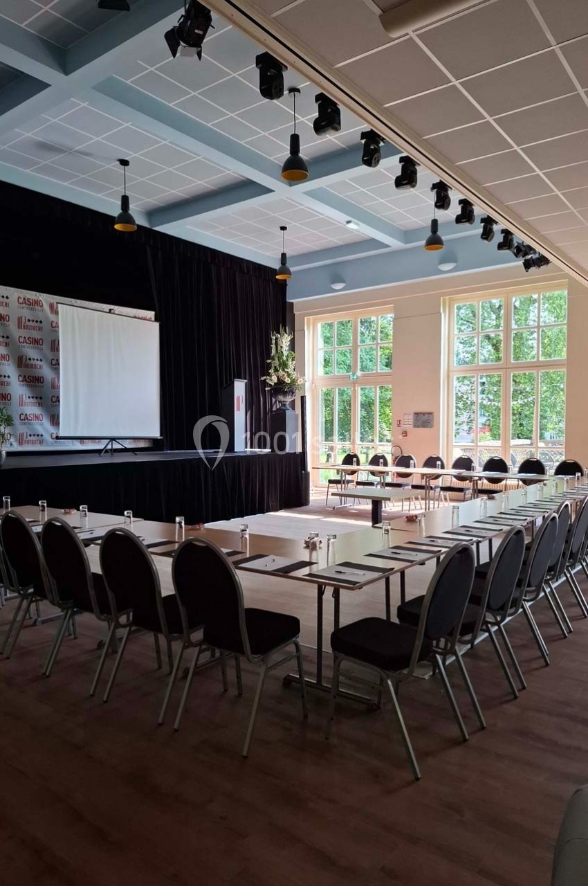 Location salle Contrexéville (Vosges) - Casino Partouche de Contrexéville #23 Salle de conférence lumineuse avec tables disposées en U, écran de projection et grandes fenêtres donnant sur l'extérieur.