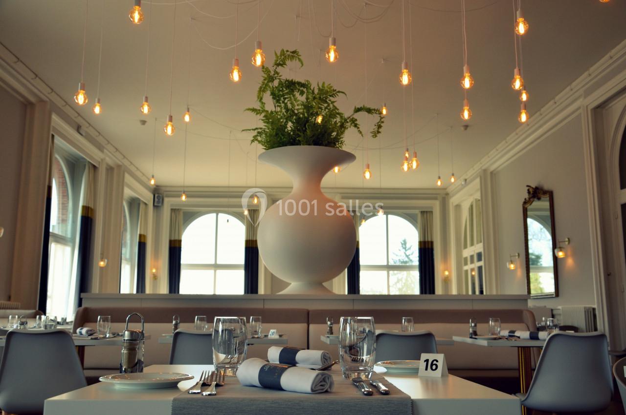 Salle de restaurant lumineuse avec tables dressées, grandes fenêtres et un vase blanc central décoré de plantes.