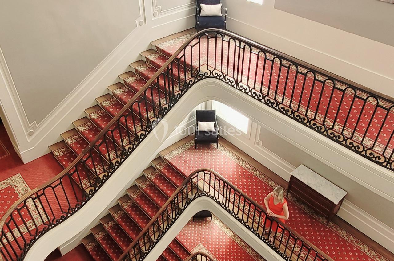 Escalier en colimaçon vu d'en haut, avec tapis rouge, rampes en fer forgé et lumière naturelle provenant des fenêtres.