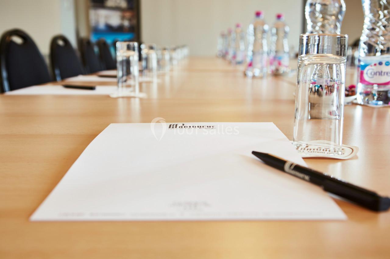Salle de réunion avec une table en bois, des blocs-notes, des stylos, des bouteilles d'eau et des verres alignés.
