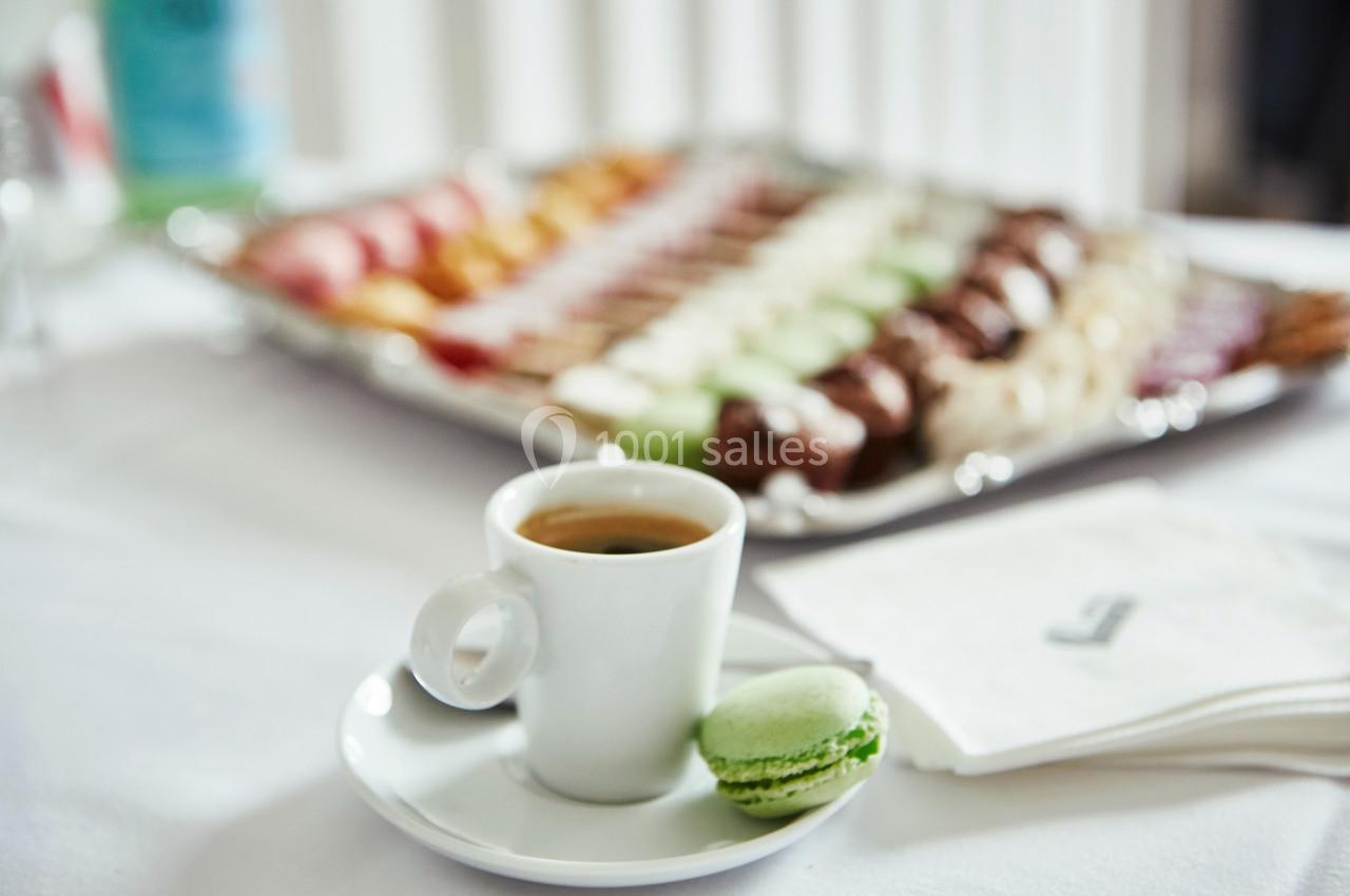 Tasse de café avec un macaron vert au premier plan, plateau de pâtisseries flou en arrière-plan.