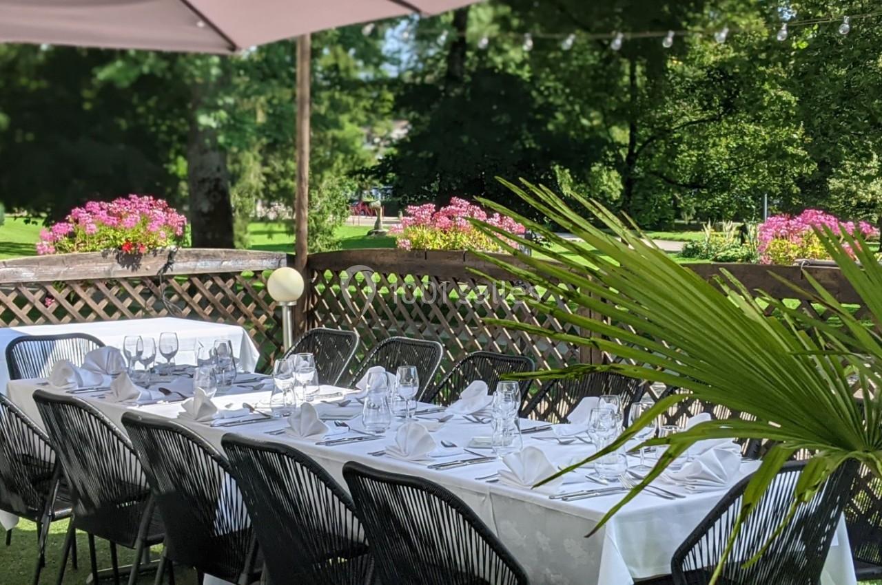 Tables dressées avec nappes blanches et chaises noires sur une terrasse ombragée, entourée de verdure et de fleurs.