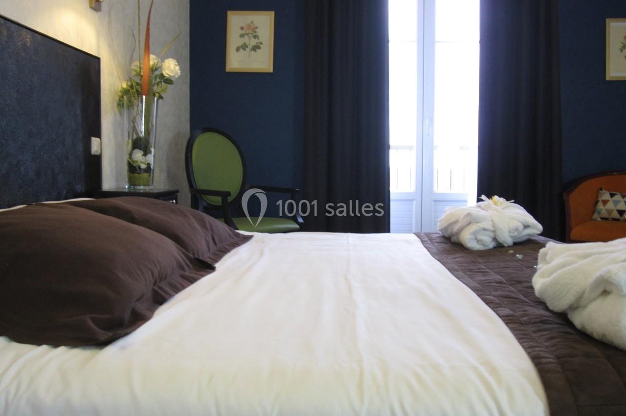 Lit double avec draps blancs et couverture marron dans une chambre lumineuse, décorée de fleurs et de cadres muraux.