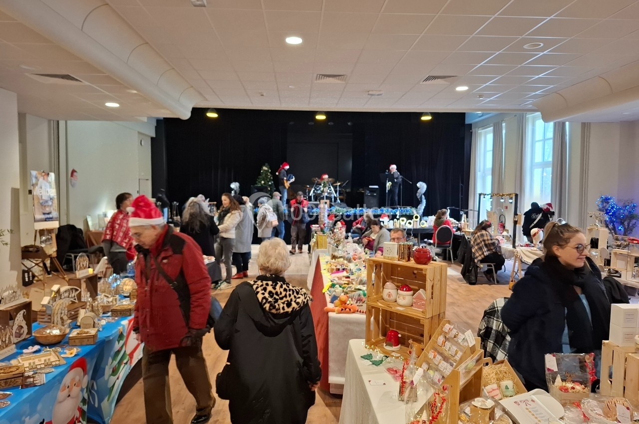 Salle décorée pour un marché de Noël avec des stands artisanaux, des visiteurs et une scène en arrière-plan.