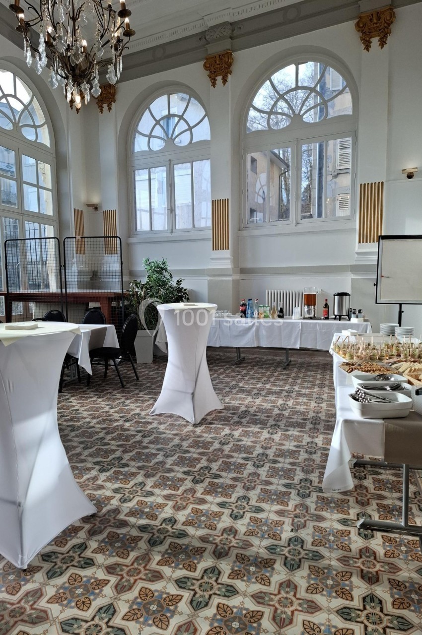 Salle lumineuse avec de grandes fenêtres, tables hautes habillées de nappes blanches et buffet disposé en arrière-plan.