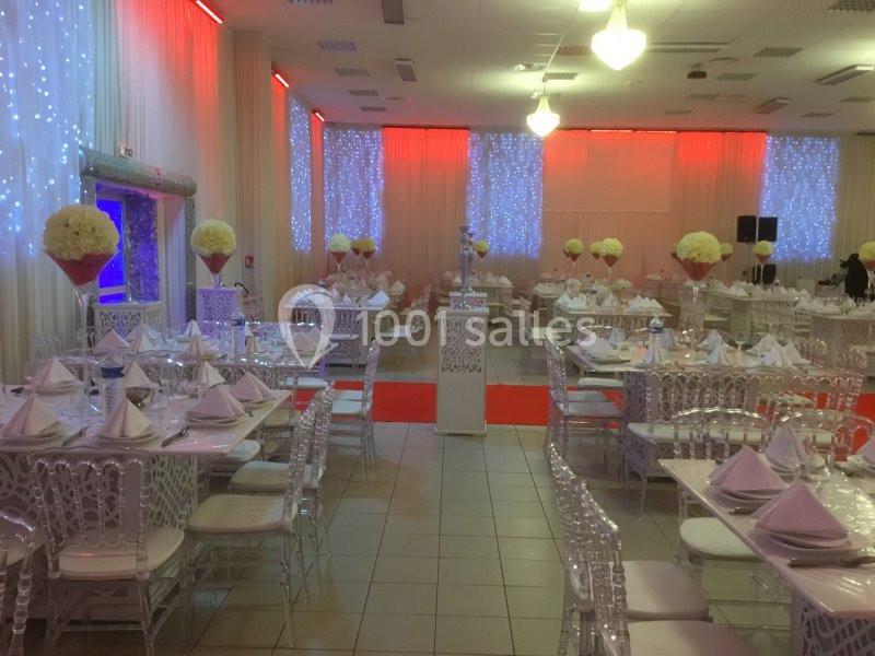 Salle de réception décorée avec des tables dressées, nappes blanches, chaises ornées et éclairage tamisé rouge et blanc.