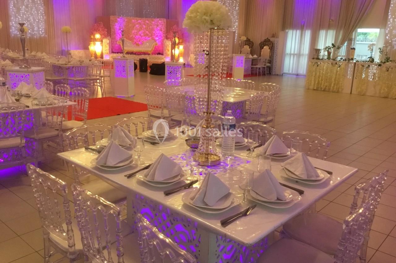 Salle de réception décorée avec des tables élégantes, nappes blanches, chaises transparentes et éclairage coloré.