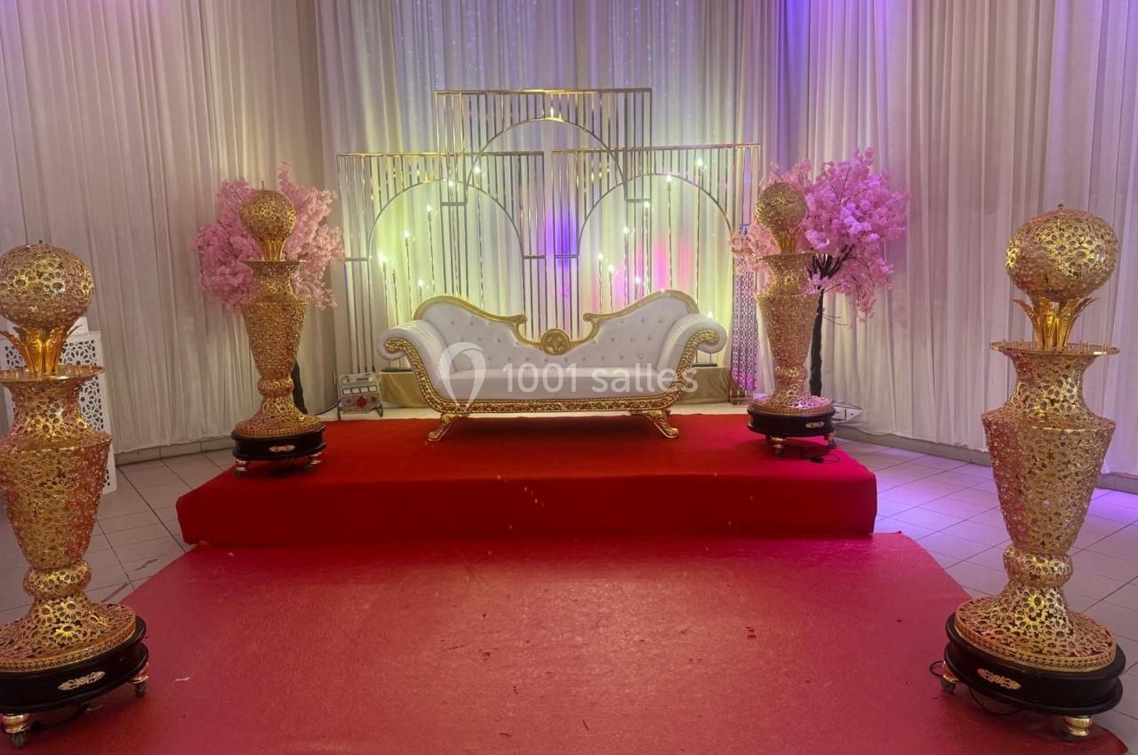 Canapé blanc orné sur une estrade rouge, entouré de colonnes dorées et de décorations florales roses.
