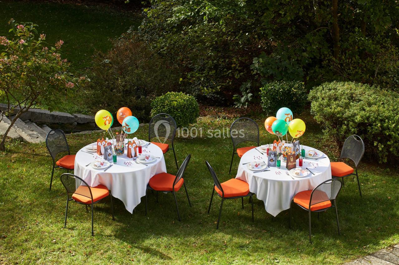 Tables rondes dressées avec nappes blanches, chaises orange, ballons et décorations, disposées dans un jardin verdoyant.