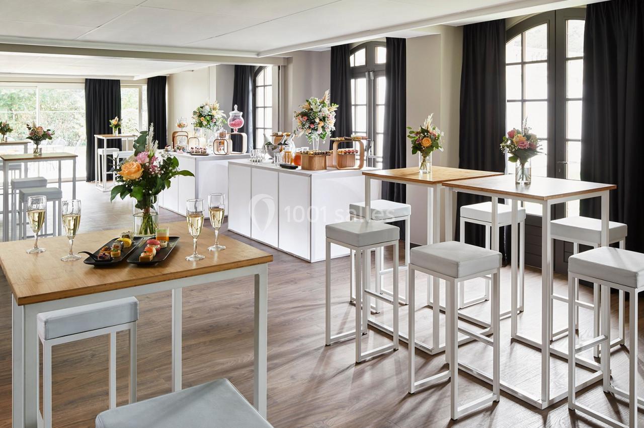 Salle lumineuse aménagée pour un événement, avec tables hautes, chaises, fleurs et buffet de boissons et amuse-bouches.