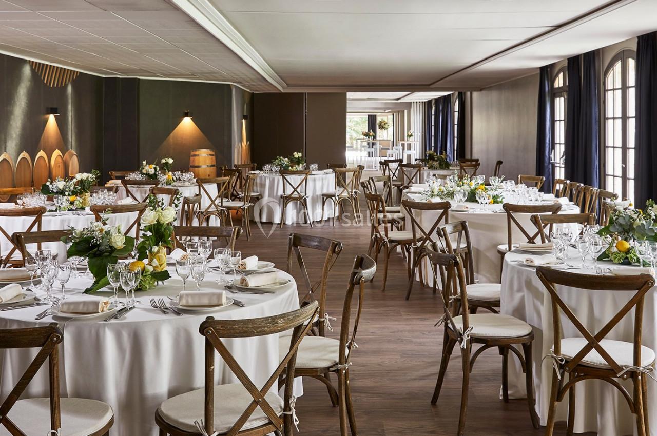 Salle de réception élégante avec tables rondes dressées, nappes blanches, chaises en bois et décor floral.
