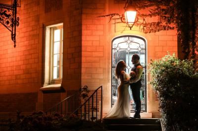 Miniature Location salle Gouvieux (Oise) - Château Jeanne & The Forest - Château de Montvillargenne #27 Un couple en tenue de mariage se tient devant une porte illuminée, entouré de briques et de végétation.