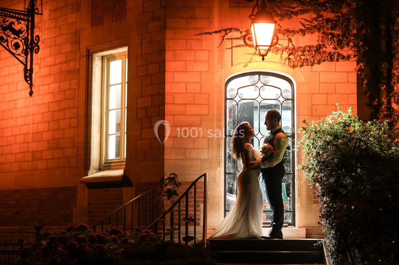 Un couple en tenue de mariage se tient devant une porte illuminée, entouré de briques et de végétation.