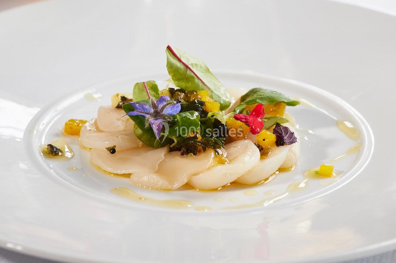 Tranches de coquilles Saint-Jacques crues garnies de fleurs comestibles, légumes colorés et feuilles vertes sur une assiette…