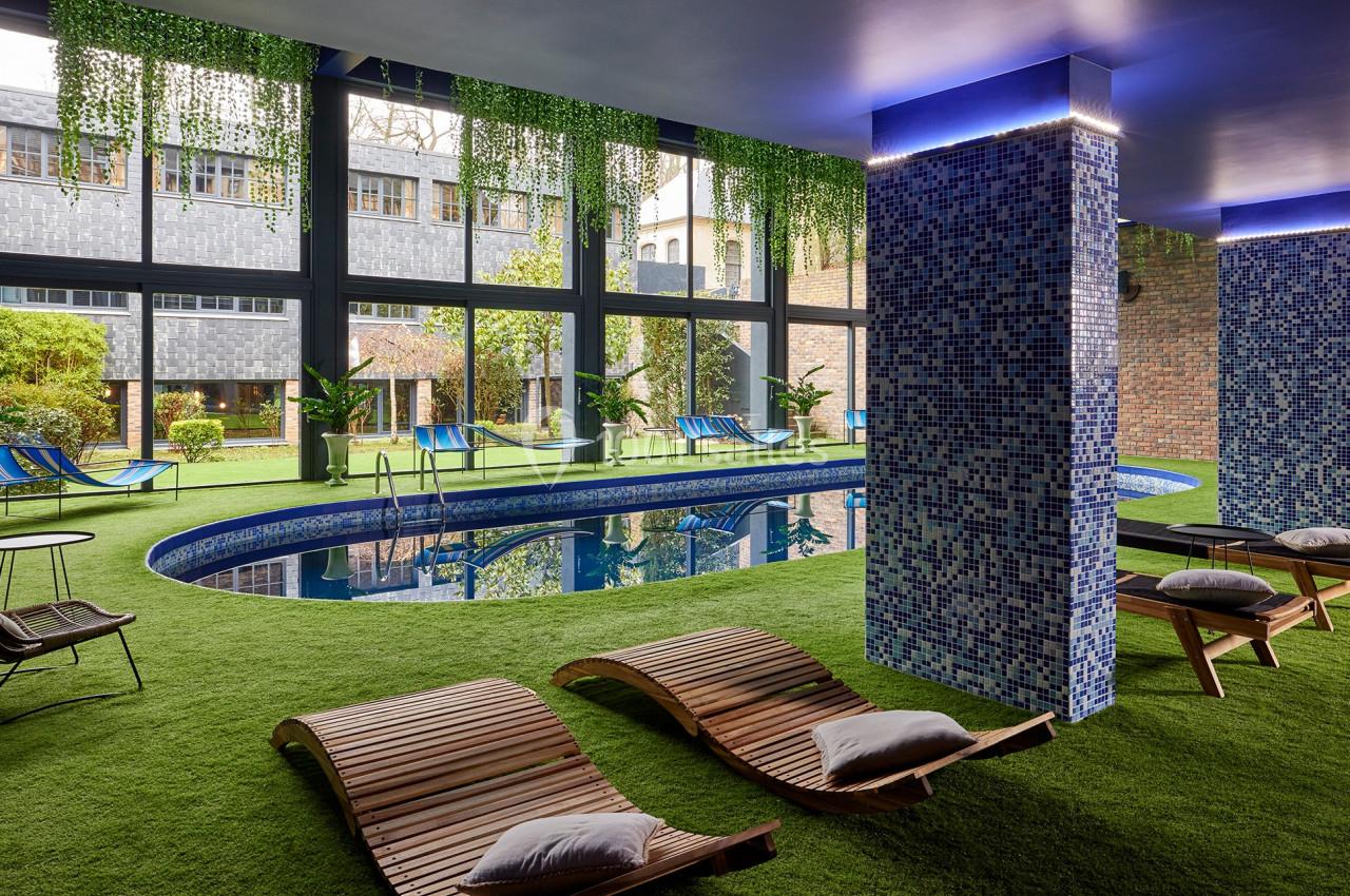 Piscine intérieure entourée de chaises longues en bois, pelouse artificielle et murs ornés de mosaïques bleues.
