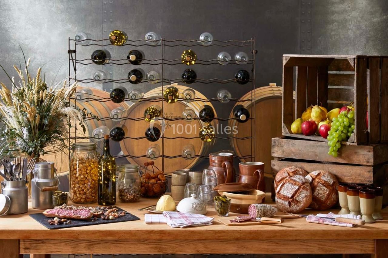 Table garnie de pains, charcuteries, fruits, pots en verre, bouteilles de vin et décor rustique avec tonneaux en arrière…