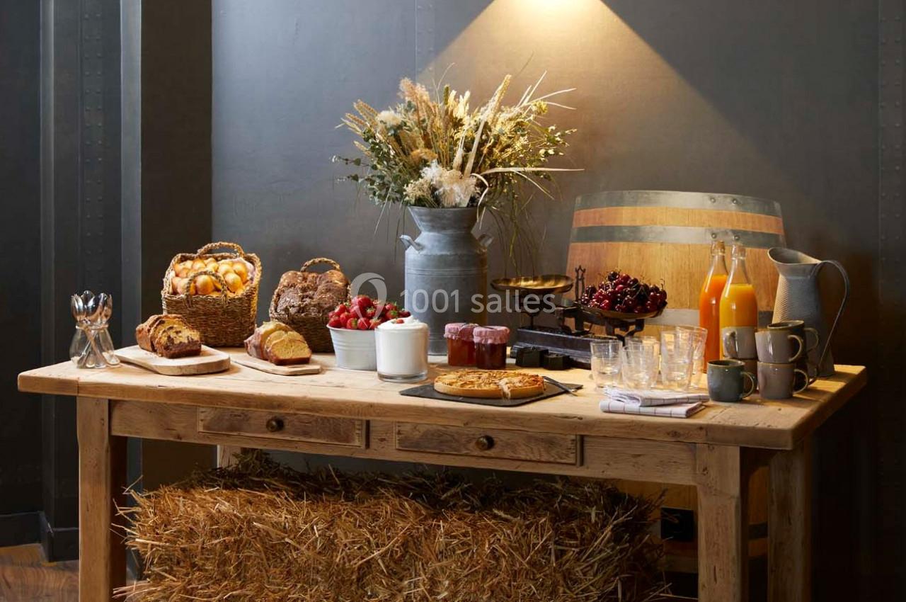 Table en bois rustique avec pains, viennoiseries, fruits, jus, confitures et fleurs séchées sous un éclairage chaleureux.