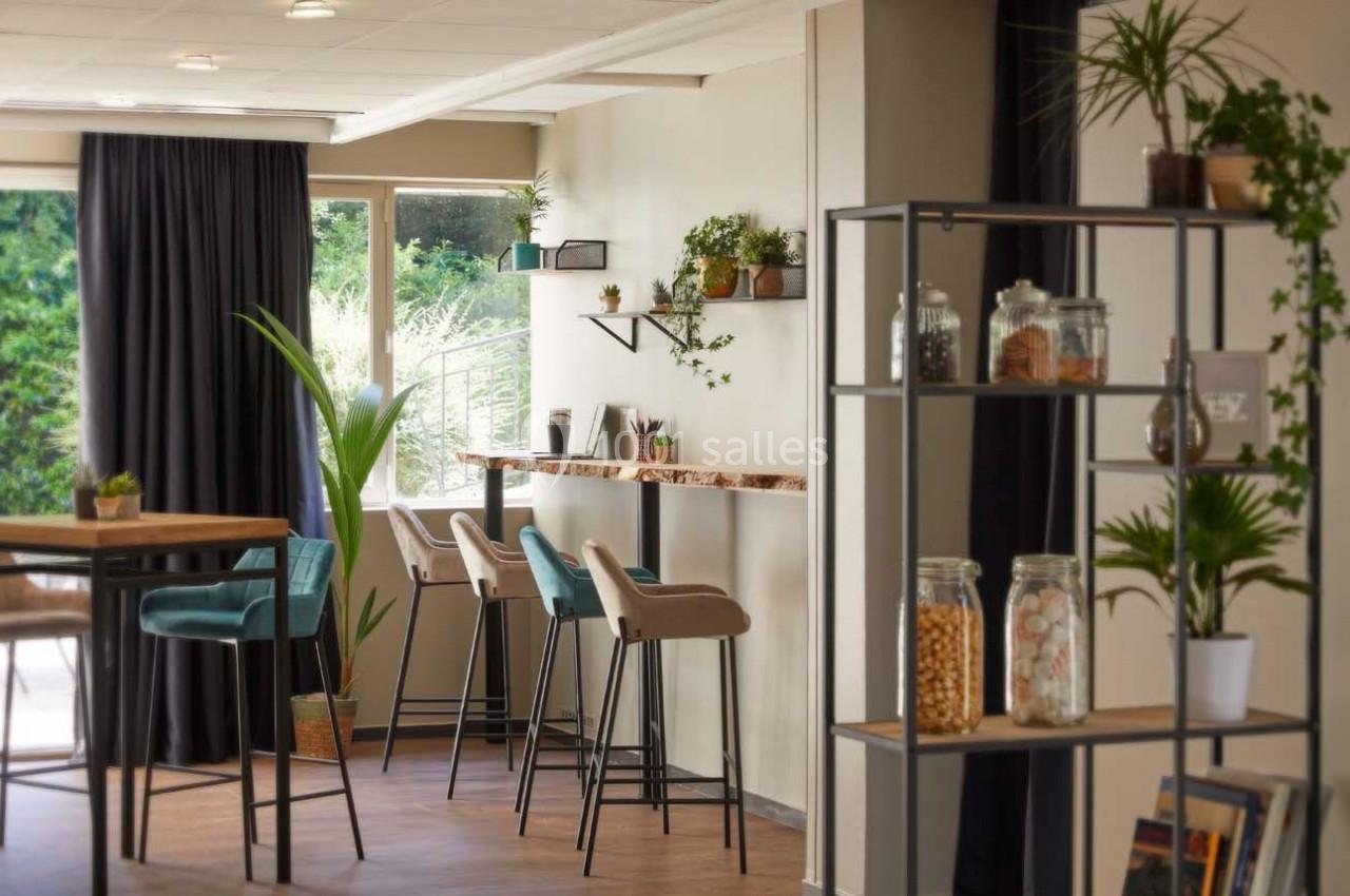 Espace lumineux avec des tabourets hauts, des étagères décorées de plantes et des bocaux, donnant sur une fenêtre.