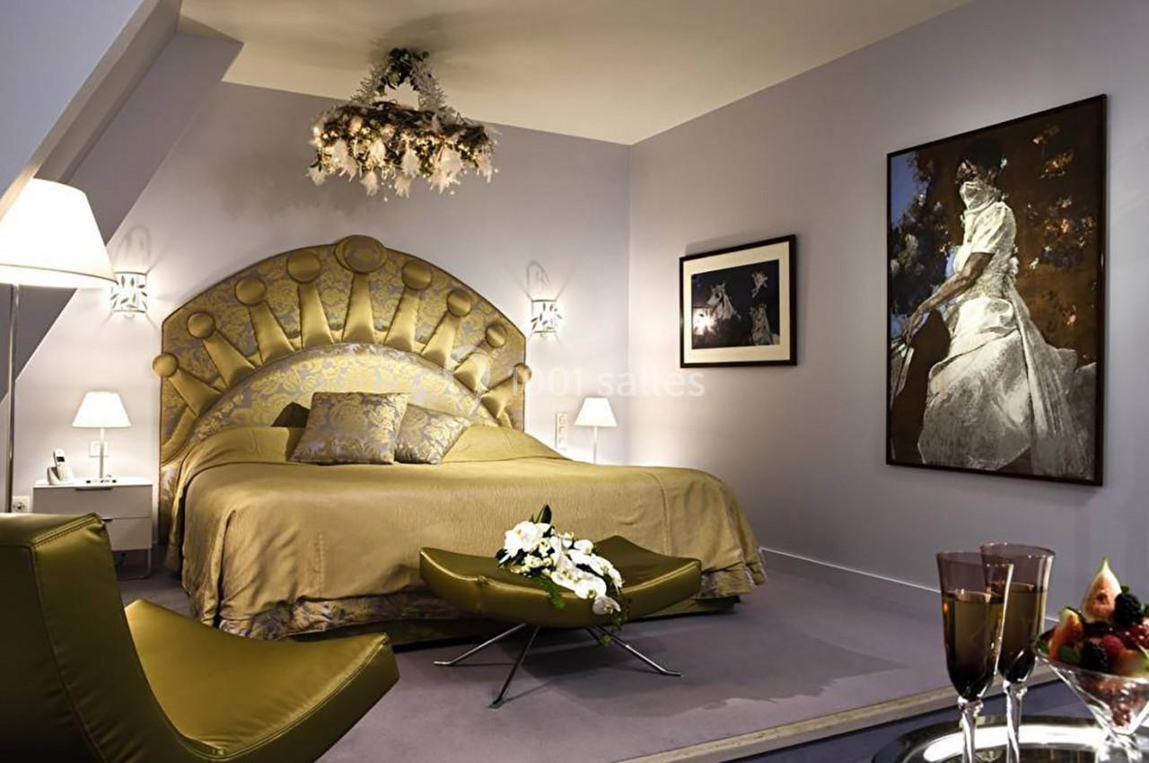 Chambre élégante avec lit double, lustre floral, fauteuil vert, tableau et éclairage tamisé.