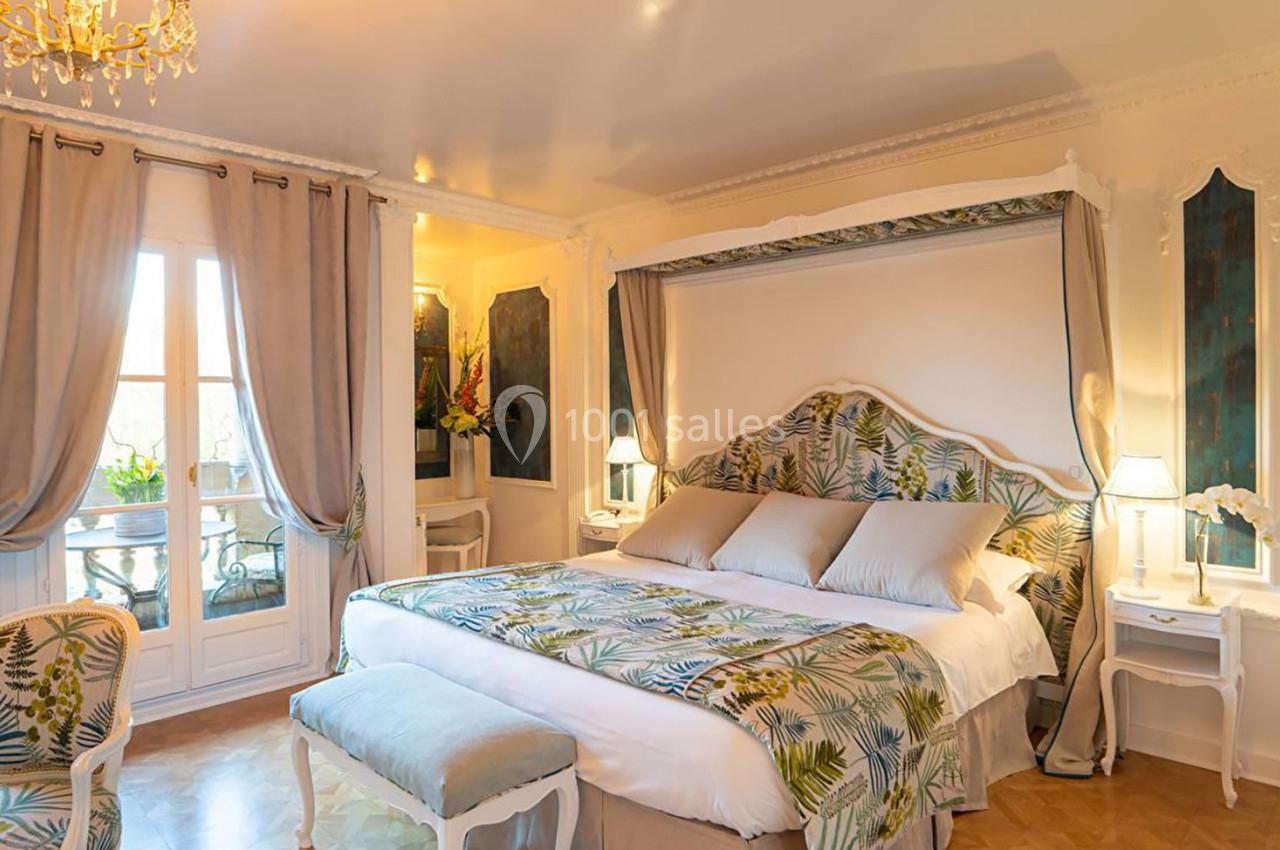 Chambre élégante avec lit double, baldaquin, décoration végétale, lumière douce et vue sur un balcon avec rideaux ouverts.