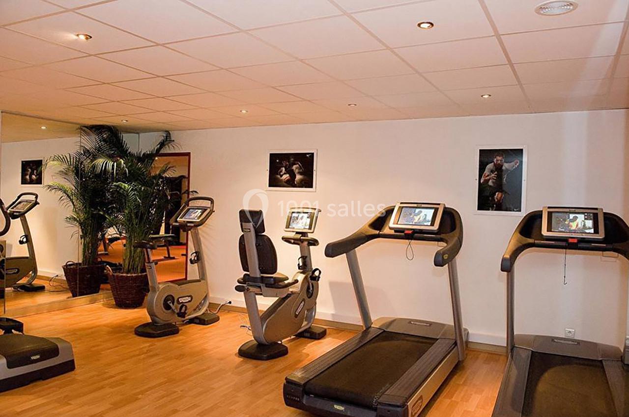 Salle de sport avec tapis de course, vélos d'exercice, plantes décoratives et affiches sur les murs.