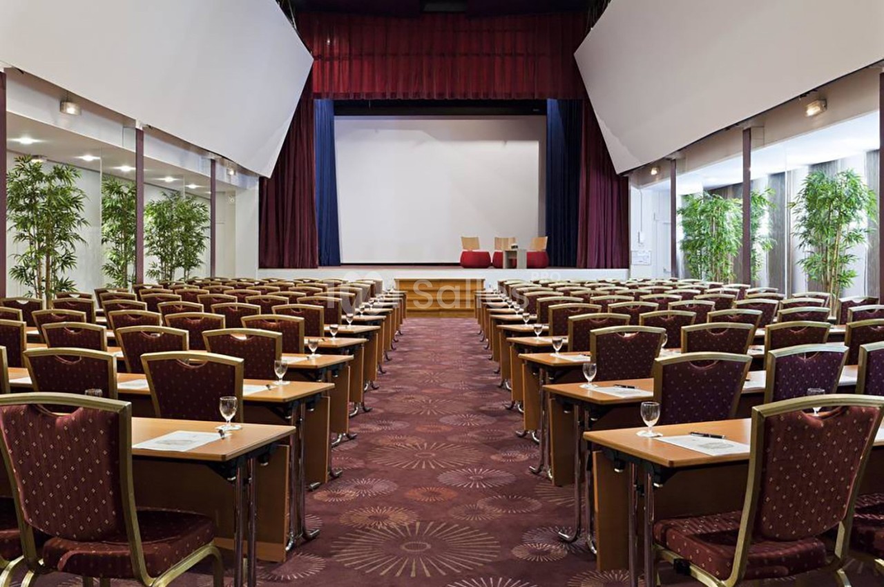 Salle de conférence avec rangées de chaises et tables alignées face à une scène équipée d'un écran blanc.
