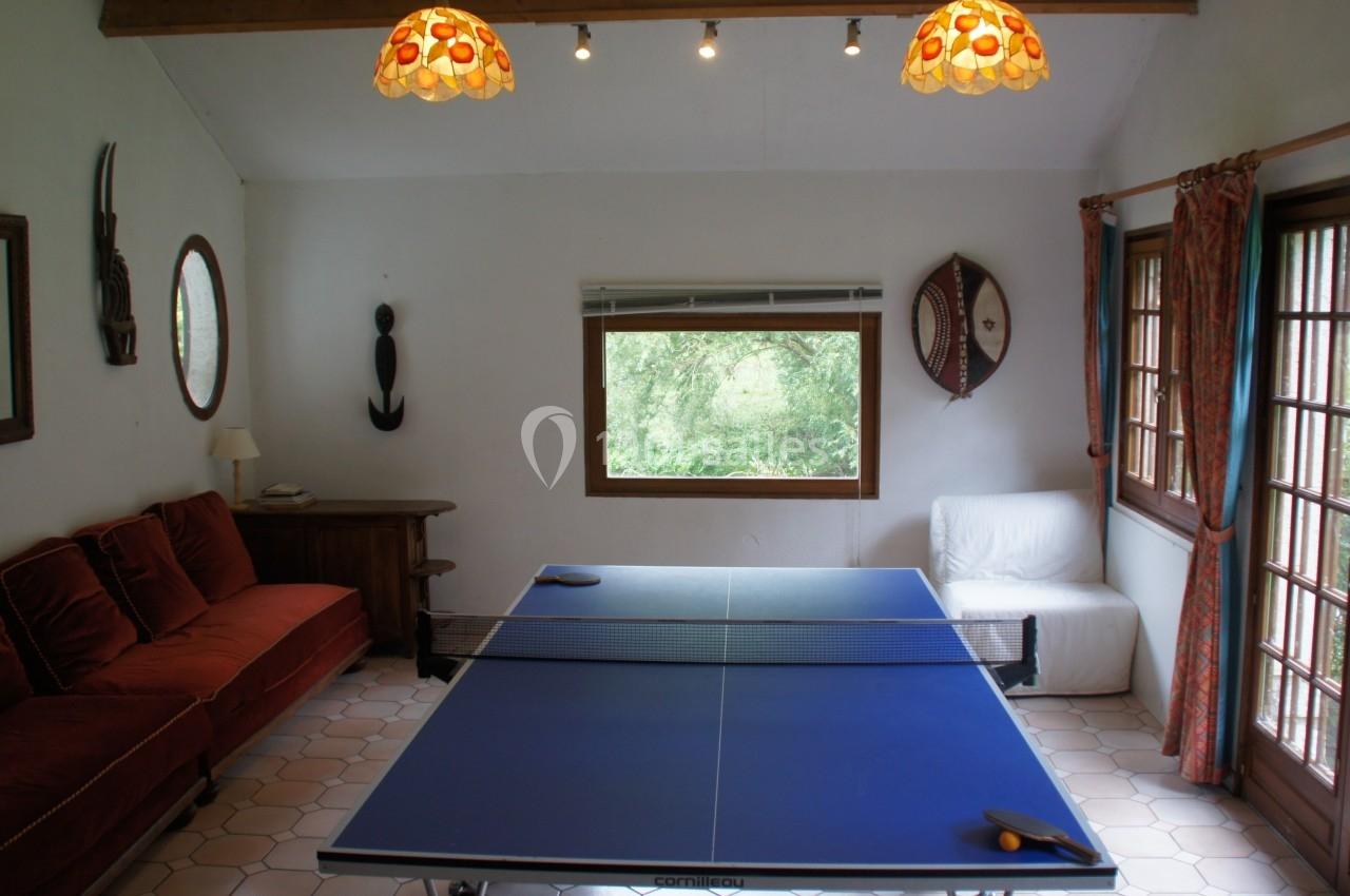 Table de ping-pong dans une pièce lumineuse avec canapé, fauteuil, décorations murales et fenêtres donnant sur la verdure.